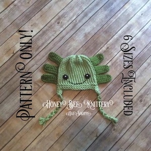 May include: Green crocheted axolotl hat with black eyes and a smiling mouth. The hat has a long tail and is on a wooden background. The text "PATTERN ONLY!" and "6 SIZES INCLUDED" is on the image.