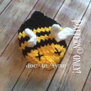May include: A handmade crochet bee-shaped item in yellow, black, and white. The bee has a black head, yellow and black striped body, white wings, and black cross-shaped eyes. The text "PATTERN ONLY!" and "HONEY BEE KNITTERY" are visible.