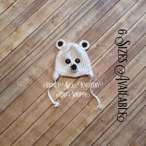 May include: White knitted polar bear hat with black eyes and nose. The hat has ear flaps and ties that can be tied under the chin. The hat is on a wooden background. 6 Sizes Available.