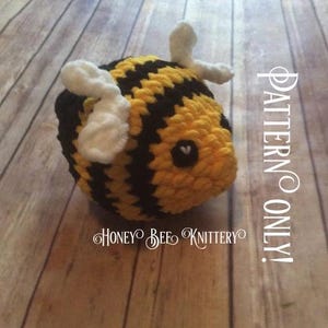 May include: A handmade, plush bee with yellow and black stripes, white wings, and a small heart-shaped face. The text "PATTERN ONLY!" and "HONEY BEE KNITTERY" are visible.
