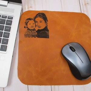 Peut inclure: Un tapis de souris en cuir marron avec une photo noir et blanc d'une mère et d'un enfant imprimée dessus. Une souris sans fil argentée est sur le tapis.