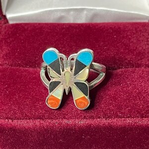 May include: A silver butterfly ring with inlaid turquoise, black, white, and red stones. The ring is in a red velvet box.