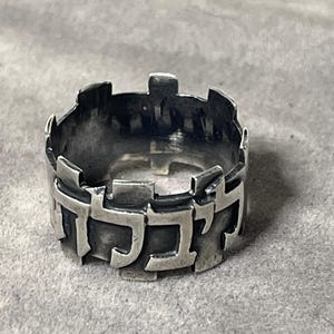 May include: A wide silver ring featuring a textured, oxidized finish and a series of raised, blocky Hebrew characters forming a continuous band around the circumference. The metal has a dark, matte appearance with sharp, geometric edges.