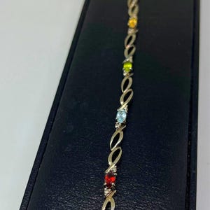 May include: A metallic bracelet featuring a series of oval-shaped links connected by small, clear stones. The design incorporates a variety of colorful, faceted gemstones in shades of red, purple, yellow, green, and light blue set in metal.