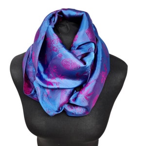 May include: A blue and fuchsia silk infinity scarf with a floral and geometric pattern. The scarf is draped over a black mannequin, highlighting its vibrant colours and intricate design. The scarf's fabric has a subtle sheen.