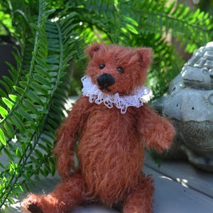 May include: A small, rust-colored teddy bear with long, shaggy fur, dark eyes, and a black nose. The bear is wearing a white, intricate lace ruff collar around its neck. It is seated in an upright position with its arms and legs extended forward.