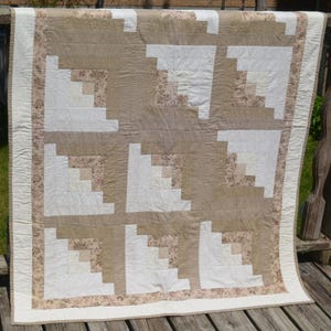 May include: A handmade quilt with a log cabin design. The quilt features squares of white, beige, and floral fabric. The border is a cream colour with a floral pattern. The quilt is displayed outdoors.