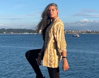 May include: A woman with long gray hair is wearing a light yellow and brown patterned shirt with long sleeves. She is standing on a pier with a body of water in the background.