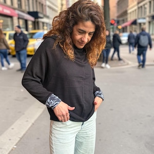 May include: A person wearing a black long-sleeve top with patterned cuffs and light blue jeans. The top has a cowl neck and the cuffs feature a white and blue wave design. The jeans are a light wash and have a wide leg.