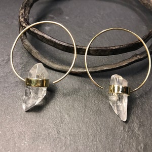 May include: A pair of gold-colored hoop earrings, each featuring a clear quartz crystal pendant. The crystals are held in place by a gold-colored band. A dark, textured object is in the background.