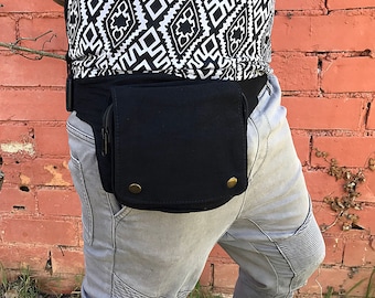 May include: A black canvas fanny pack with two zippered pockets and two gold-toned buttons. The fanny pack is worn around the waist of a person wearing gray jeans.