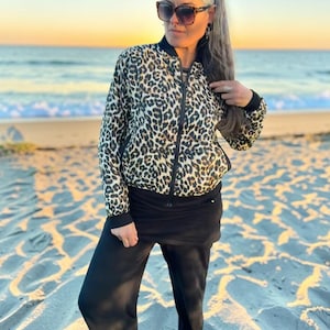 Puede incluir: Una chaqueta bomber con estampado de leopardo con ribete negro y cierre de cremallera. La chaqueta se combina con pantalones negros. El fondo muestra una escena de playa con arena y el océano.