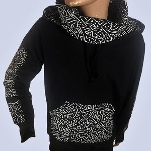 May include: Black hoodie with a white geometric pattern on the hood, sleeves, and front pocket. The hoodie has a drawstring and long sleeves. The design is a repeating pattern of lines and shapes.