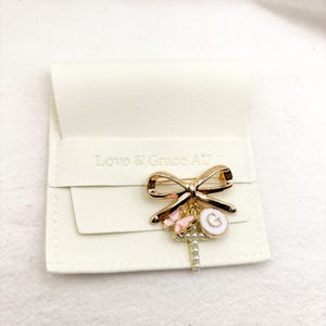 May include: Gold bow brooch with a charm of a cross, a butterfly, and a letter G. The brooch is on a white velvet background. Love & Grace AU is printed on the background.