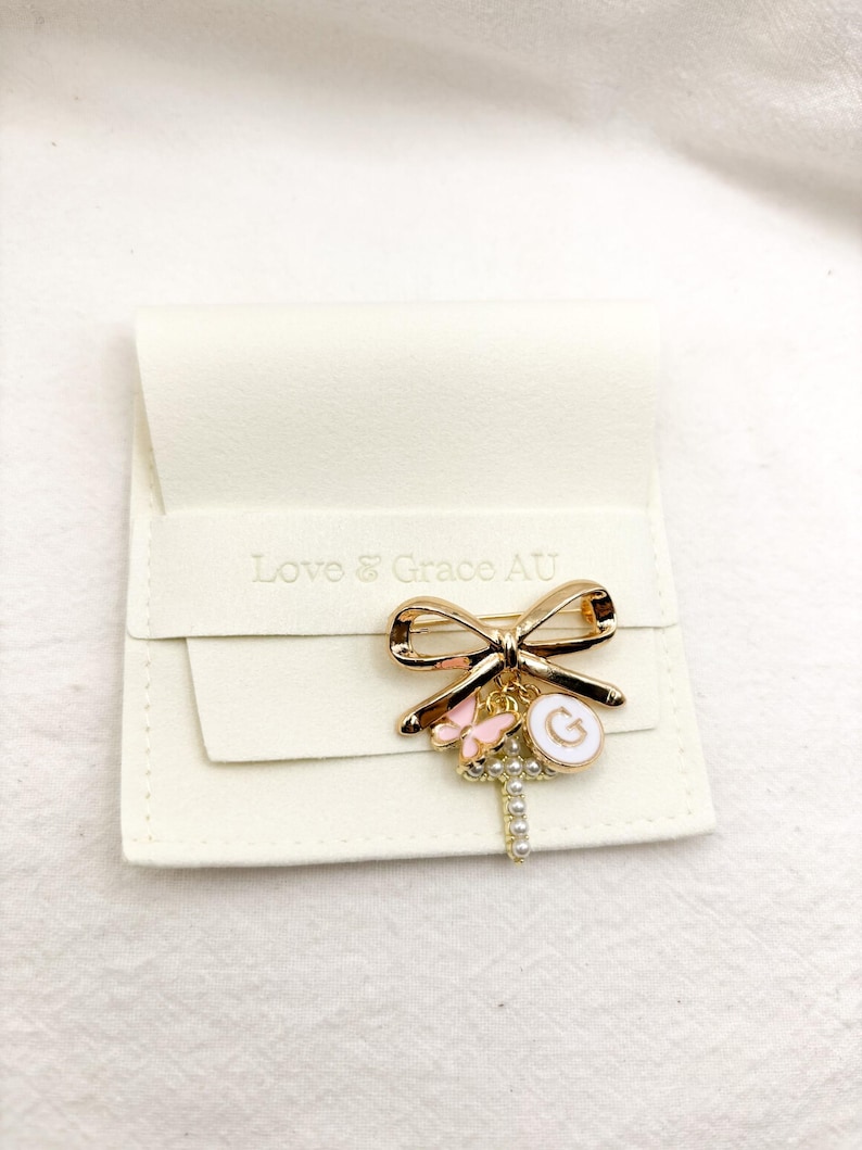 May include: Gold bow brooch with a charm of a cross, a butterfly, and a letter G. The brooch is on a white velvet background. Love & Grace AU is printed on the background.