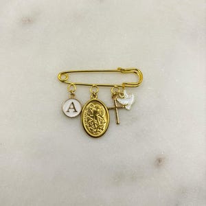 May include: Gold-tone safety pin with multiple charms. Charms include a white enamel circle with the letter "A", a gold oval pendant with a religious figure, a gold cross, and a white dove. The pin is displayed on a white marble surface.