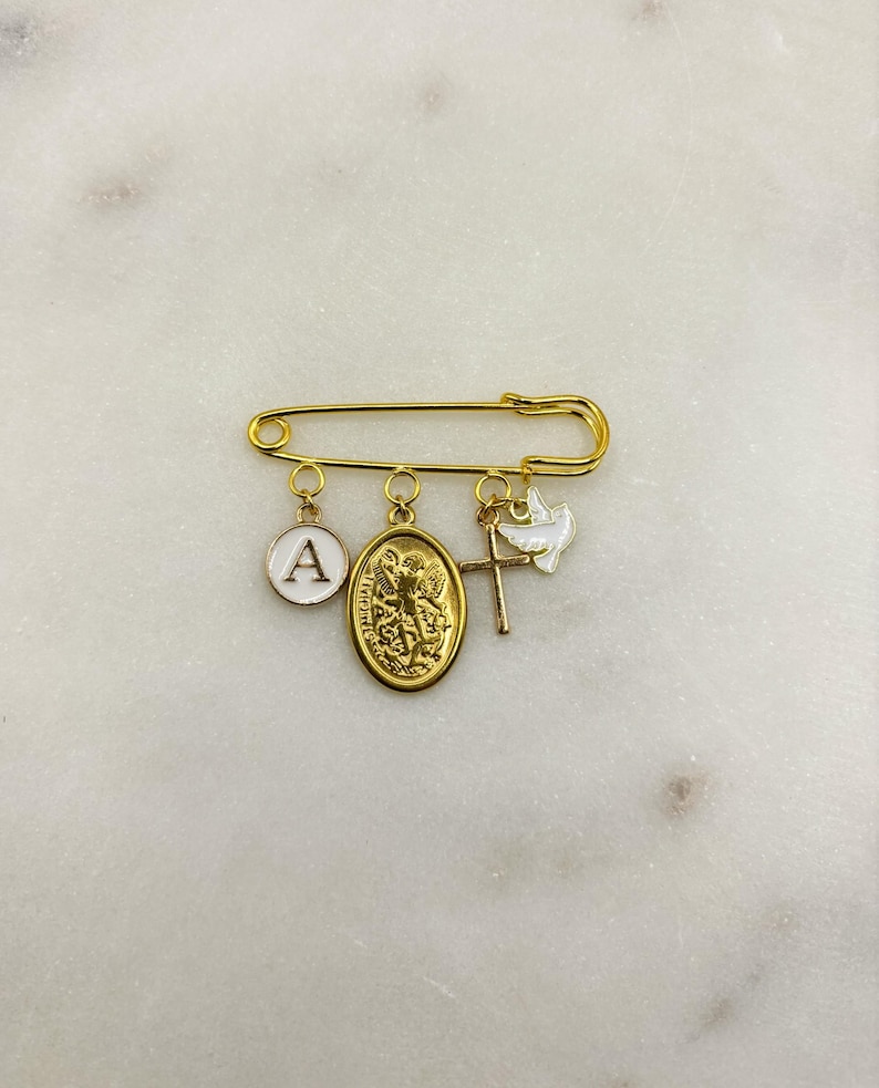 May include: Gold-tone safety pin with multiple charms. Charms include a white enamel circle with the letter "A", a gold oval pendant with a religious figure, a gold cross, and a white dove. The pin is displayed on a white marble surface.