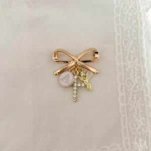 May include: Gold-tone bow brooch with a charm of a letter A, a cherub, and a cross with rhinestones.