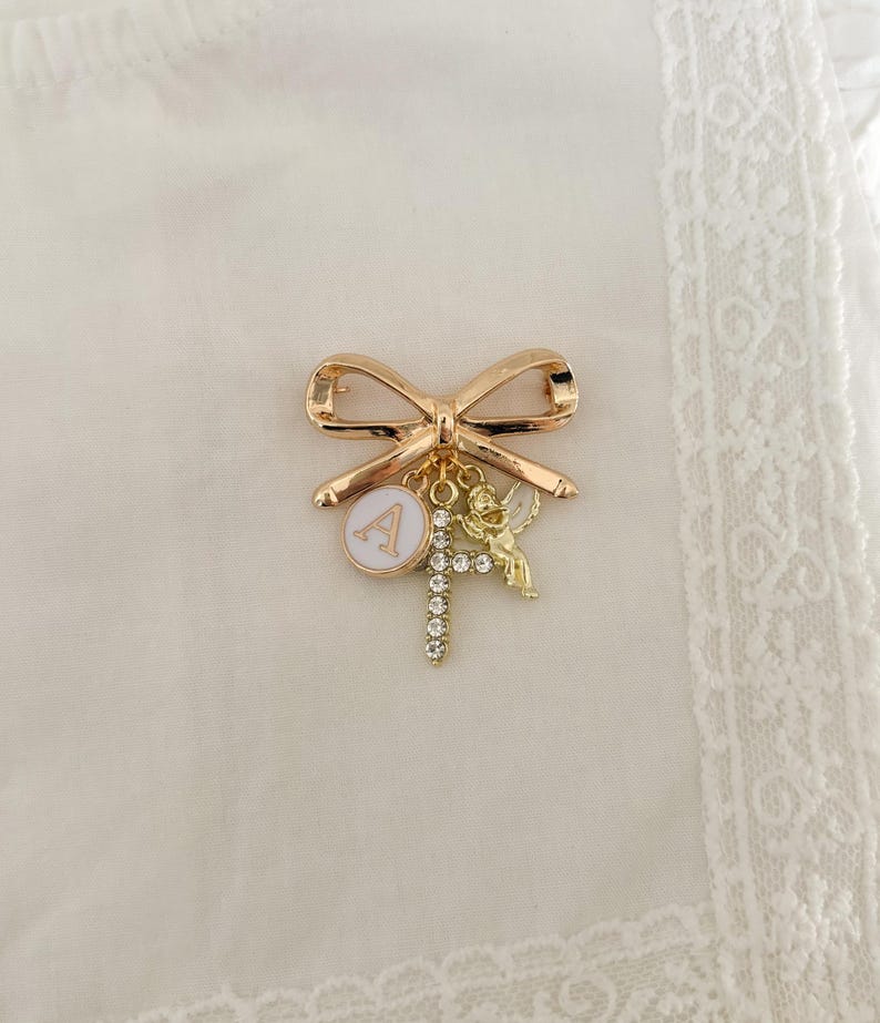 May include: Gold-tone bow brooch with a charm of a letter A, a cherub, and a cross with rhinestones.