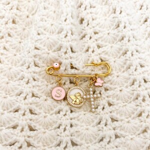 May include: A gold-tone safety pin brooch featuring a small pink bead, a circular charm with the letter S, a circular charm with a gold angel figure, a cross charm with white beads, and a small pink butterfly charm hanging from the pin.