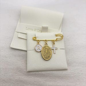 May include: A gold-colored safety pin with three charms: a white circle with the letter "R", an oval medallion, and a white cross. The pin is displayed on a white surface, with a white pouch in the background. The pouch has the text "Love & Grace".