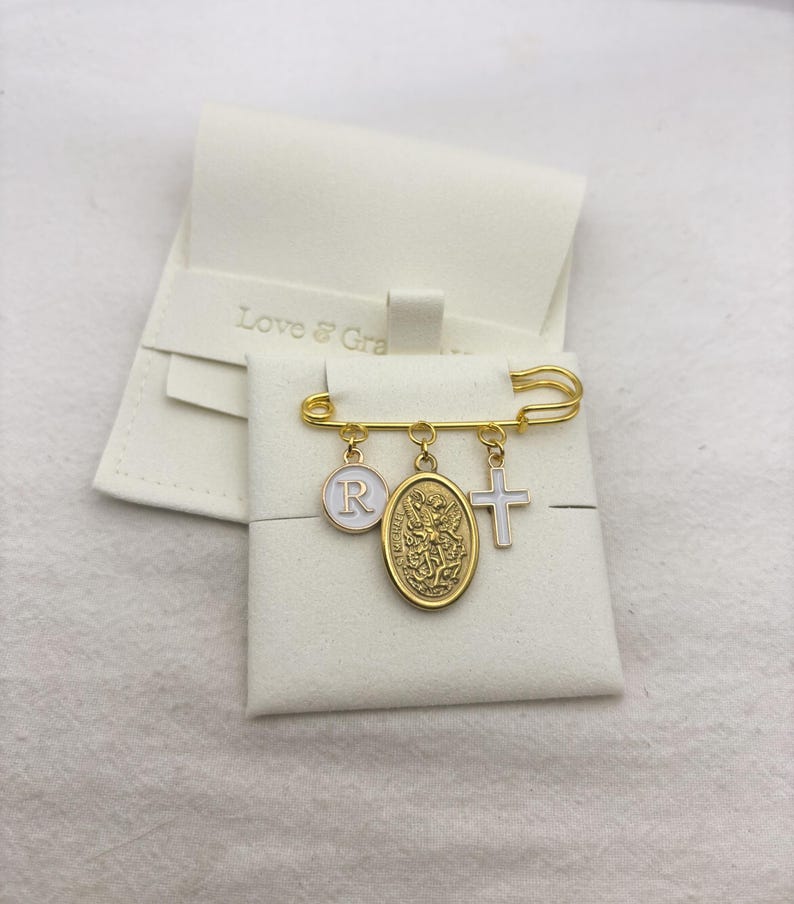May include: A gold-colored safety pin with three charms: a white circle with the letter "R", an oval medallion, and a white cross. The pin is displayed on a white surface, with a white pouch in the background. The pouch has the text "Love & Grace".