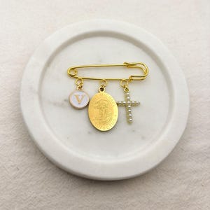 May include: Gold-tone safety pin brooch with three charms: a white enamel circle with a "V", an oval Saint Michael medal, and a pearl-studded cross. The brooch is displayed on a white, circular tray.