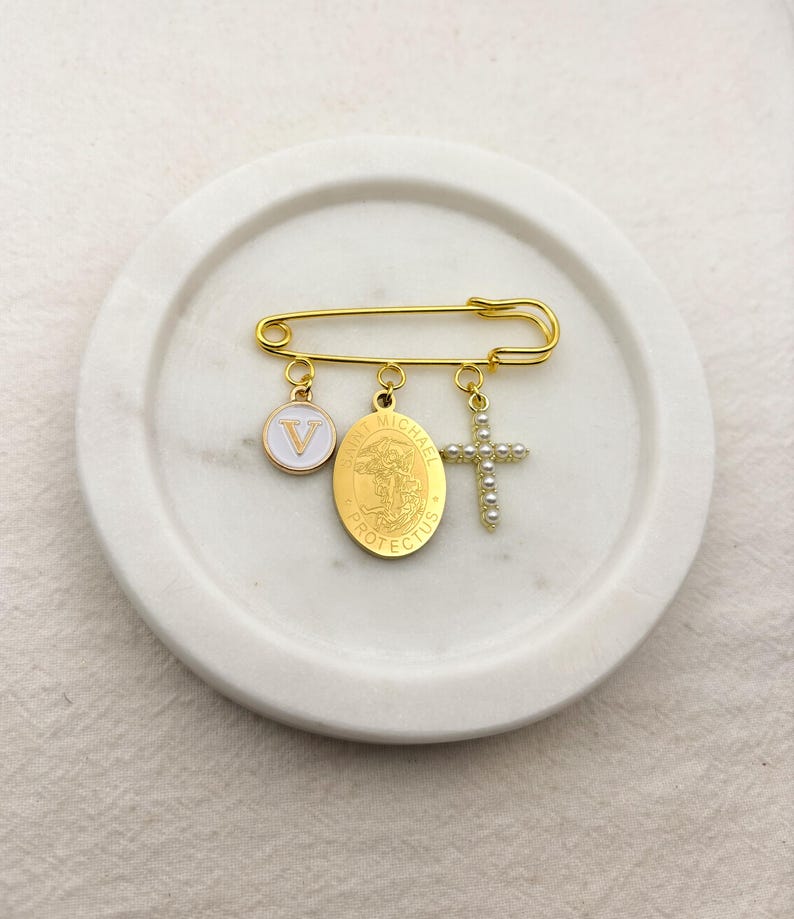 May include: Gold-tone safety pin brooch with three charms: a white enamel circle with a "V", an oval Saint Michael medal, and a pearl-studded cross. The brooch is displayed on a white, circular tray.
