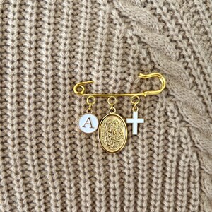 May include: Gold-colored safety pin with three charms: a white circle with the letter "A", a gold medallion, and a white cross. The pin is displayed on a textured, tan fabric background.