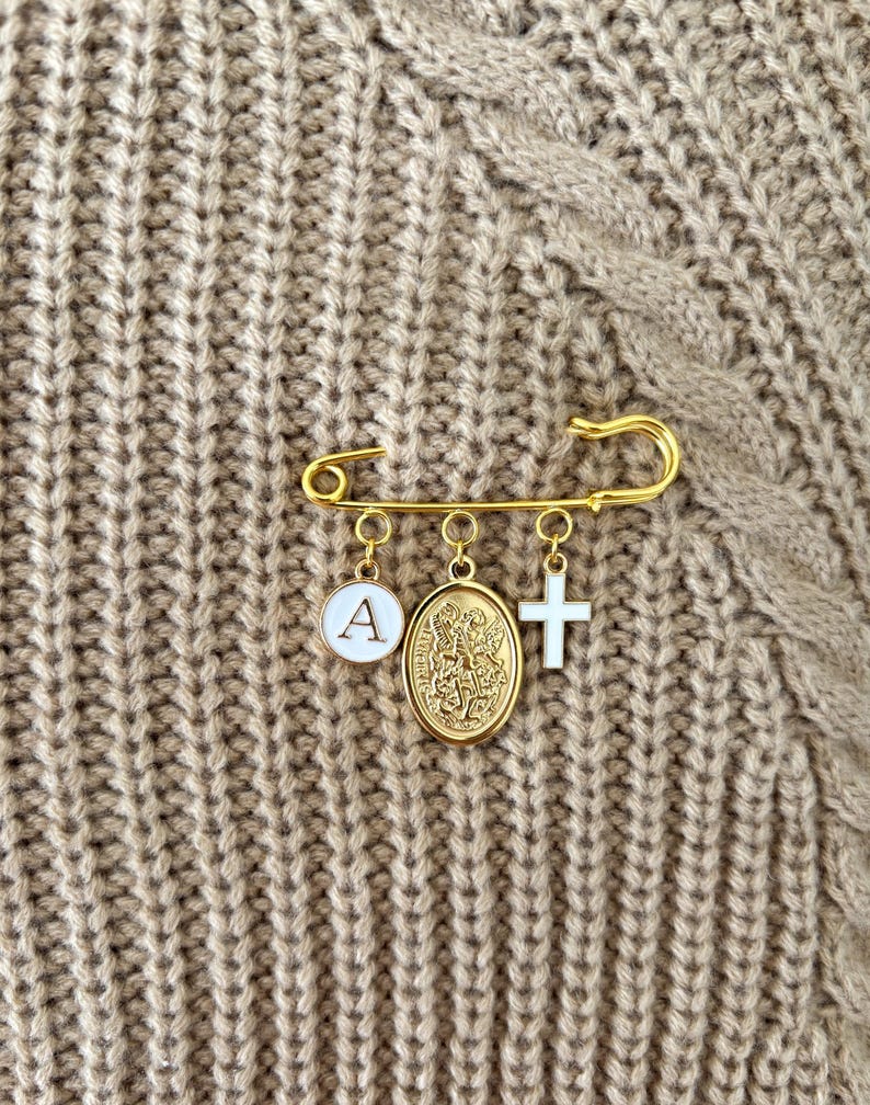 May include: Gold-colored safety pin with three charms: a white circle with the letter "A", a gold medallion, and a white cross. The pin is displayed on a textured, tan fabric background.