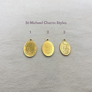 May include: Three gold-colored Saint Michael charm pendants. The pendants are oval-shaped with a loop at the top for hanging. The text "St Michael Charm Styles" is above the pendants. Each pendant has a different design.