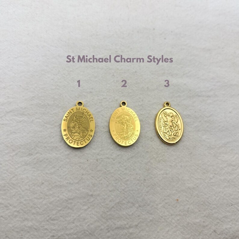 May include: Three gold-colored Saint Michael charm pendants. The pendants are oval-shaped with a loop at the top for hanging. The text "St Michael Charm Styles" is above the pendants. Each pendant has a different design.