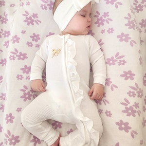 May include: A white baby onesie with a ruffle down the front and a gold bow detail. The onesie is worn by a baby with a white headband and bow on a white and purple floral blanket.