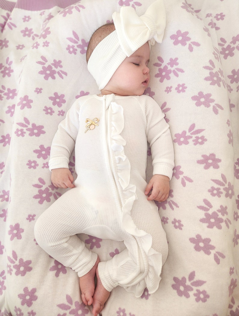 May include: A white baby onesie with a ruffle down the front and a gold bow detail. The onesie is worn by a baby with a white headband and bow on a white and purple floral blanket.