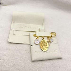 May include: Gold-tone safety pin with multiple charms, including a white and gold letter "V" pendant, a Saint Michael medal, and a cross with a dove. The pin is displayed on a white fabric with the text "Love & Grace AU".