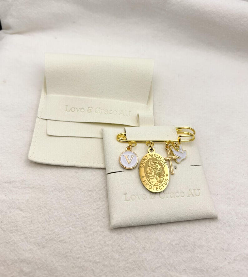 May include: Gold-tone safety pin with multiple charms, including a white and gold letter "V" pendant, a Saint Michael medal, and a cross with a dove. The pin is displayed on a white fabric with the text "Love & Grace AU".