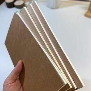 May include: A set of three rectangular notebooks with plain brown covers and white paper pages. The notebooks are bound with visible white thread stitching along the spine, showing a simple construction with clean edges and a neutral color palette.