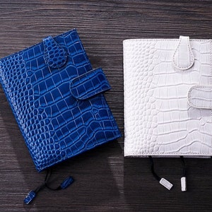 K&ouml;nnte beinhalten: Zwei Lederjournale mit Krokodilhautstruktur. Ein Journal ist leuchtend blau, das andere wei&szlig;. Beide haben einen Klappenverschluss und ein passendes Lesezeichen. Die Journale werden auf einer dunklen Holzoberfl&auml;che pr&auml;sentiert.