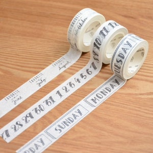 Peut inclure: Trois rouleaux de ruban adhésif décoratif avec du texte noir sur fond blanc. Le ruban présente des dates, des mois et des jours de la semaine. Le ruban est déroulé sur une surface en bois.