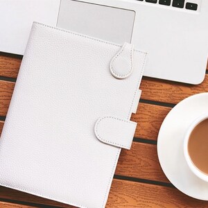 K&ouml;nnte beinhalten: Ein wei&szlig;er Lederplaner mit Tab-Verschluss liegt offen auf einem Holztisch neben einem Laptop und einer Kaffeetasse. Der Planer hat eine strukturierte Oberfl&auml;che und einen Schlaufenverschluss. Eine wei&szlig;e Untertasse enth&auml;lt eine Tasse Kaffee.