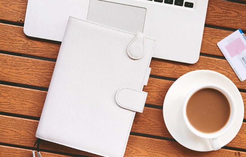 K&ouml;nnte beinhalten: Ein wei&szlig;er Lederplaner mit Tab-Verschluss liegt offen auf einem Holztisch neben einem Laptop und einer Kaffeetasse. Der Planer hat eine strukturierte Oberfl&auml;che und einen Schlaufenverschluss. Eine wei&szlig;e Untertasse enth&auml;lt eine Tasse Kaffee.