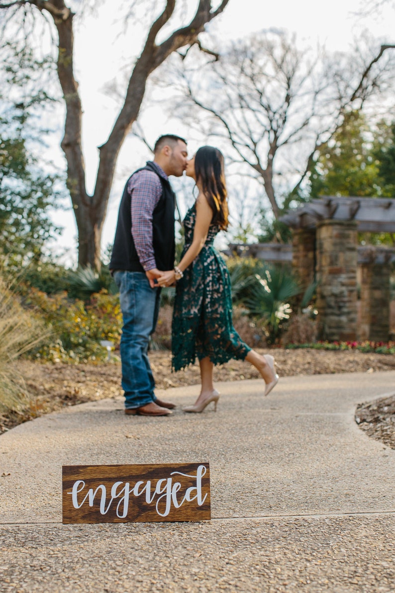May include: A couple in love kisses in front of a wooden sign that reads "engaged" on a pathway in a garden.