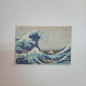 Könnte beinhalten: Ein rechteckiger Druck von Hokusais "Die große Welle vor Kanagawa". Das Kunstwerk zeigt eine große Meereswelle mit Booten und dem Berg Fuji im Hintergrund. Der Druck hat einen gezackten Rand und Text in der oberen linken Ecke. Die Welle ist blau und weiß.