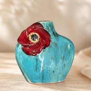 May include: A turquoise ceramic vase with a glossy finish and a dark, mottled texture. A large, three-dimensional dark red poppy flower with a textured yellow and black centre is attached to the upper left side of the vase body.