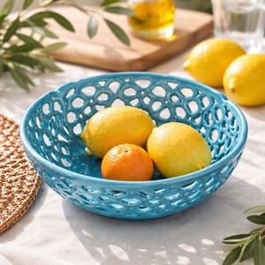 Może przedstawiać: Jasnoturkusowa ceramiczna miska o skomplikowanym, ażurowym wzorze siatki. W misce znajdują się dwie żółte cytryny i jeden pomarańczowy owoc cytrusowy, co tworzy żywy kontrast między niebieskim naczyniem a świeżymi owocami wewnątrz.