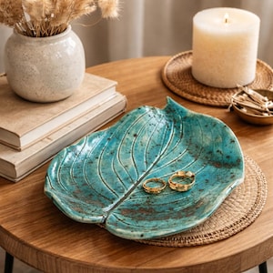 Può includere: Un piatto a forma di foglia in ceramica turchese contiene due anelli d'oro. Il piatto è appoggiato su un sottobicchiere rotondo e intrecciato su un tavolo di legno. Una candela accesa, un piccolo vaso con fiori secchi e libri sono anche sul tavolo.