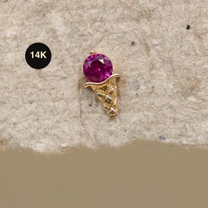 Puede incluir: Un pendiente de botón con forma de cono de helado dorado con una gema redonda de color magenta. El pendiente está detallado con un diseño de cono de gofre y una pequeña etiqueta redonda negra que dice "14K".