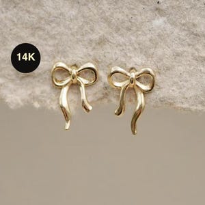 May include: A pair of gold-coloured bow-shaped earrings. The earrings are small and delicate, with a polished finish. The image includes a black circle with the text "14K".