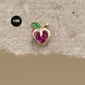 Pode incluir: Um brinco dourado em forma de pêssego. O pêssego tem um centro em forma de coração, na cor magenta, e uma folha verde. O brinco é carimbado com "14K".