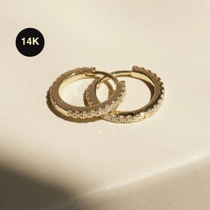 May include: Two gold 14K hoop earrings with small, clear stones set around the outer edge. The earrings are displayed on a cream-colored surface. Text on the image reads, "Sold as a Single" and "14K".